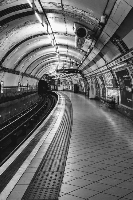 Interior view of Swiss Cottage underground station showing a curved platform with white tiled walls adorned with advertisements, including one for a sleep aid device, and a large London Underground ro